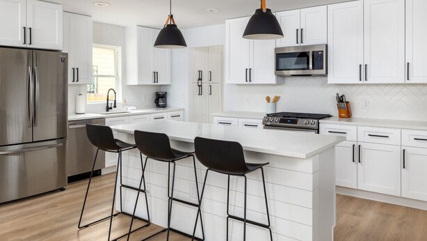 Een moderne keuken met witte kastjes, een roestvrijstalen koelkast, een magnetron en een gasfornuis. Er staan drie zwarte barkrukken bij het eiland.