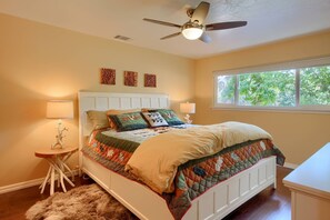 A bedroom with a bed, two bedside lamps, a ceiling fan, a window with a view of trees, and a small table with a lamp.