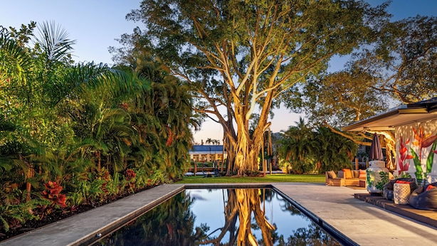 Una piscina con el reflejo de árboles, rodeada de abundante vegetación y mobiliario exterior.