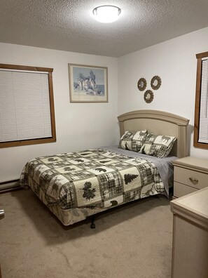 Een slaapkamer met een bed, nachtkastje, dressoir, raam met jaloezieën, plafondlamp en een ingelijst schilderij aan de muur.