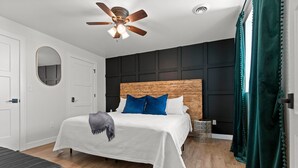 A bedroom with a wooden headboard, a bed with white bedding and blue pillows, a ceiling fan, and a dark green curtain.