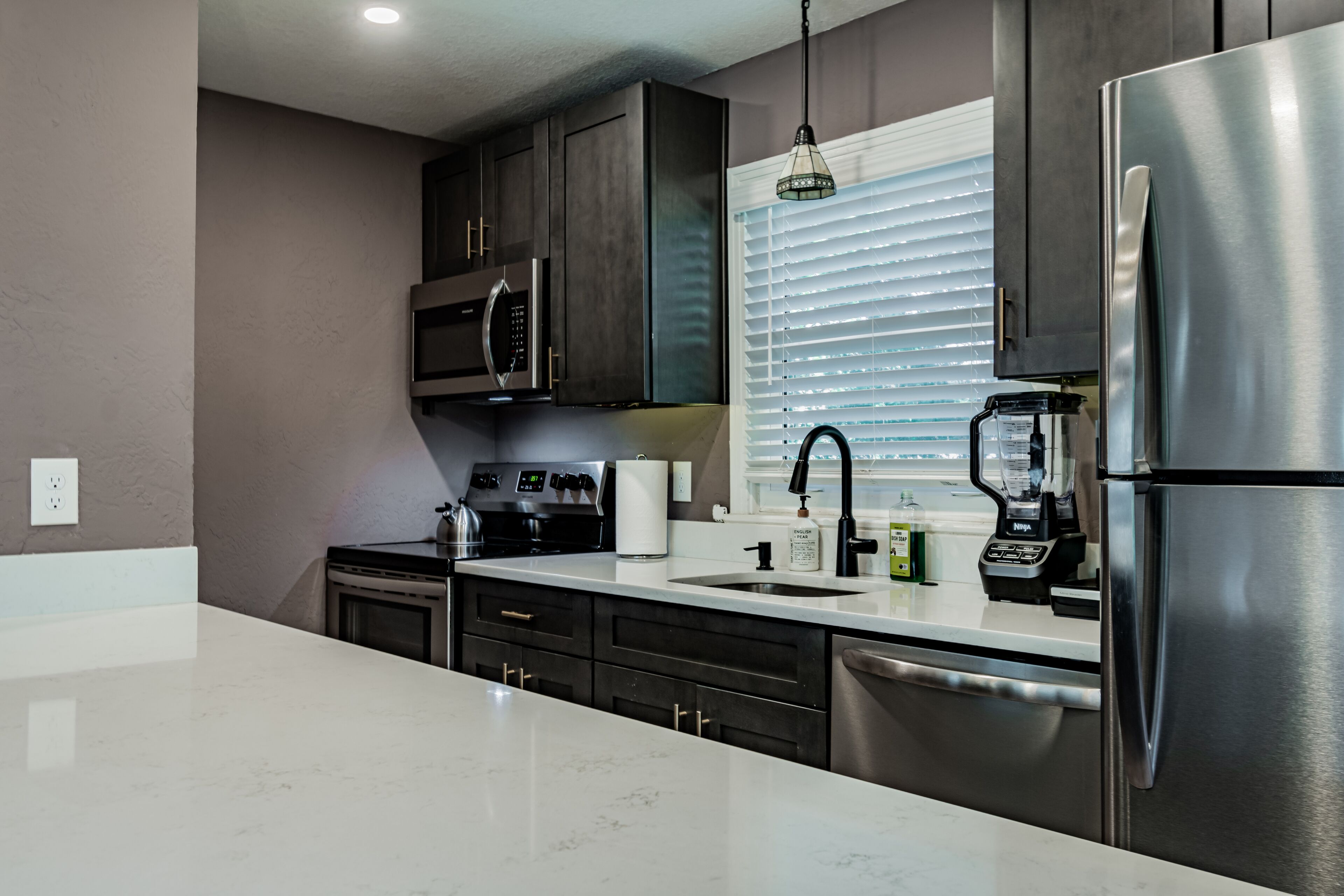 A modern kitchen with dark cabinets, stainless steel appliances, a white countertop, and a window with blinds.