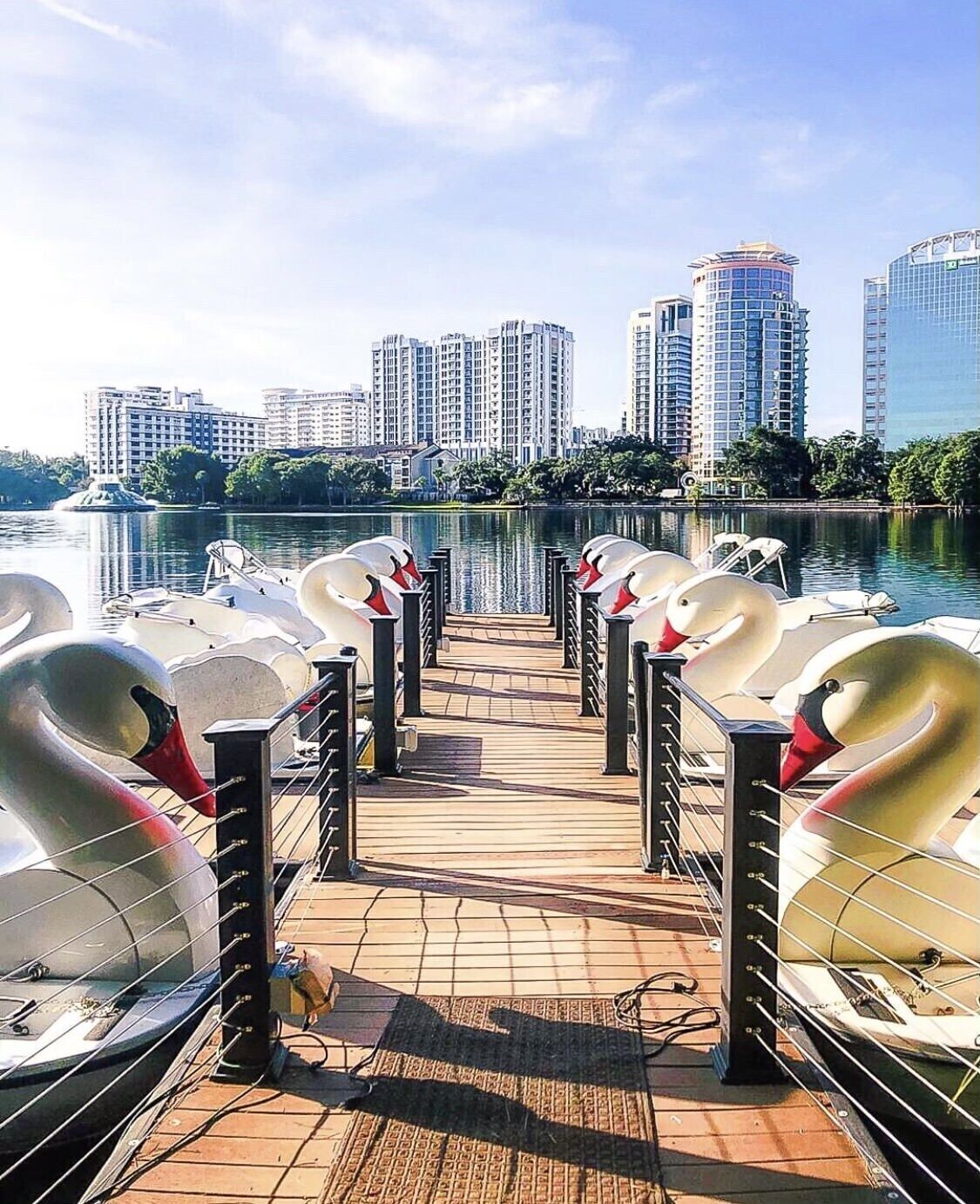 A pier with swan-shaped boats docked, leading to a body of water with a cityscape in the background.