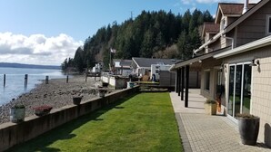 Ein Ufergrundstück mit Rasenfläche, Kübelpflanzen und Blick auf das Wasser sowie bewaldete Hügel.