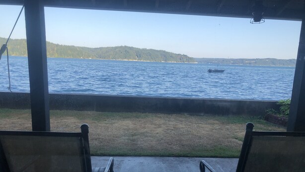 Blick von einer überdachten Sitzgelegenheit mit zwei Stühlen auf ein Gewässer, eine Wiese und eine entfernte Küste.