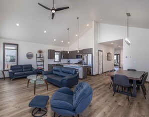 A modern living room with a sofa, armchair, and ottoman, a dining area with a table and chairs, and a kitchen with a refrigerator and microwave.
