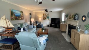A living room with a sofa, armchair, coffee table, and a fireplace. There is a television, a ceiling fan, and a painting on the wall. - Plantation Styled Home with Sunroom and Large Deck/Yard (Augusta)