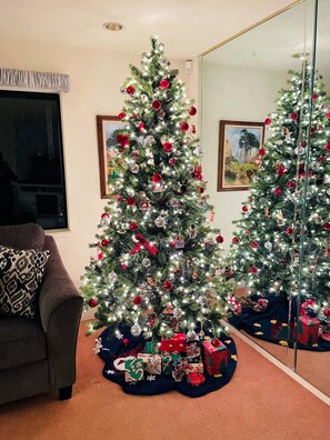 A living room with a Christmas tree decorated with ornaments and lights, a couch with a patterned pillow, and a large mirror reflecting the tree.