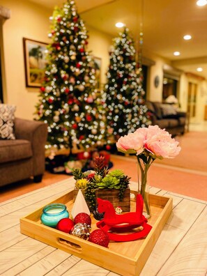 Ein Holztablett mit einer Kerze, roten und silbernen Verzierungen und einer rosa Blume in einer Vase, auf einem Holzboden, im Hintergrund ein Weihnachtsbaum.