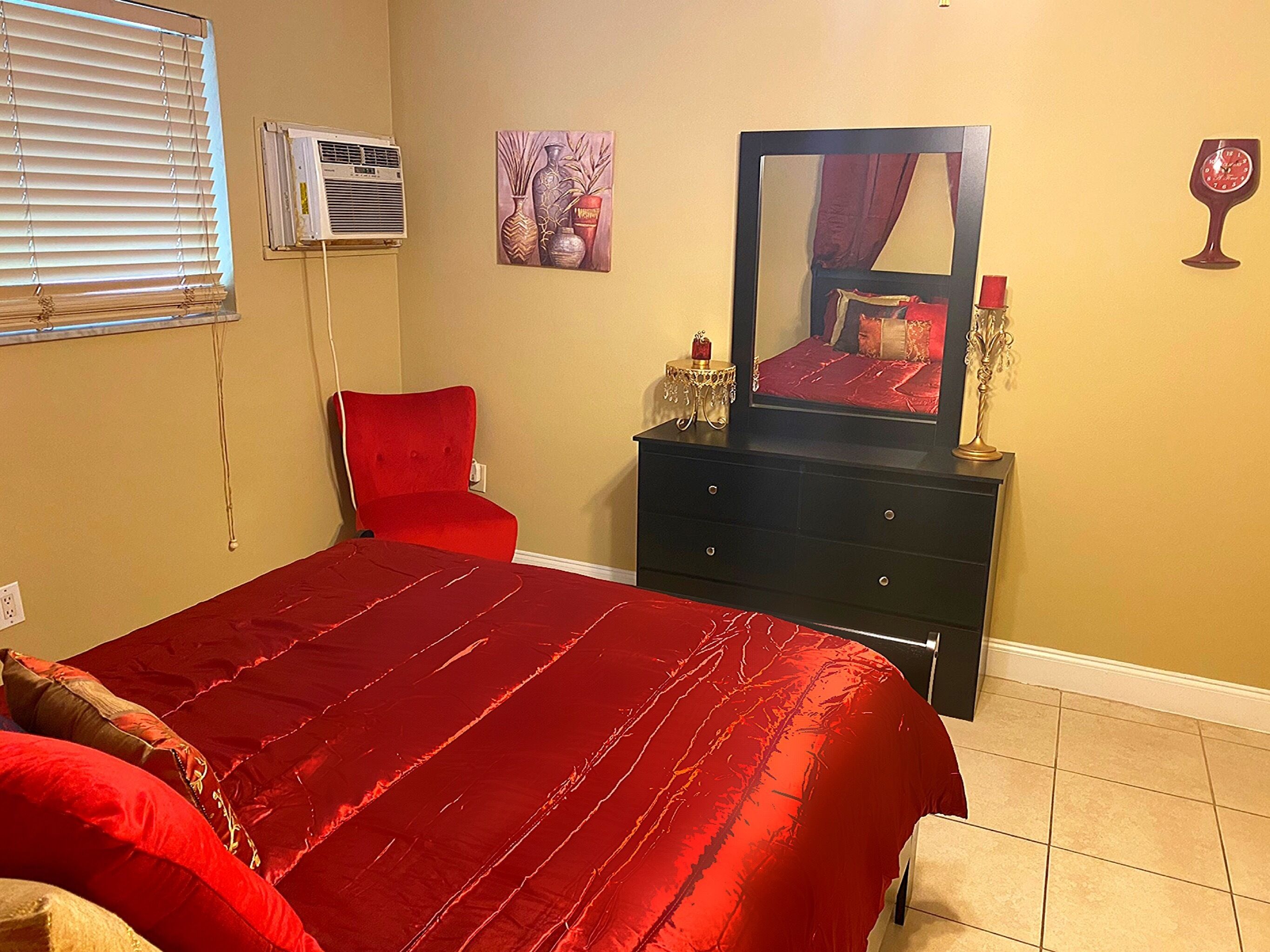 A bedroom with a red bedspread, a red chair, a dresser with a mirror, and a wall-mounted air conditioner.