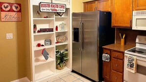 Una cocina con un refrigerador de acero inoxidable, armarios de madera y un microondas. Hay carteles en la pared, incluyendo uno que dice 'ROOSTER ROASTER COFFEE' y otro que dice 'KITCHEN NEVER CLOSED'.