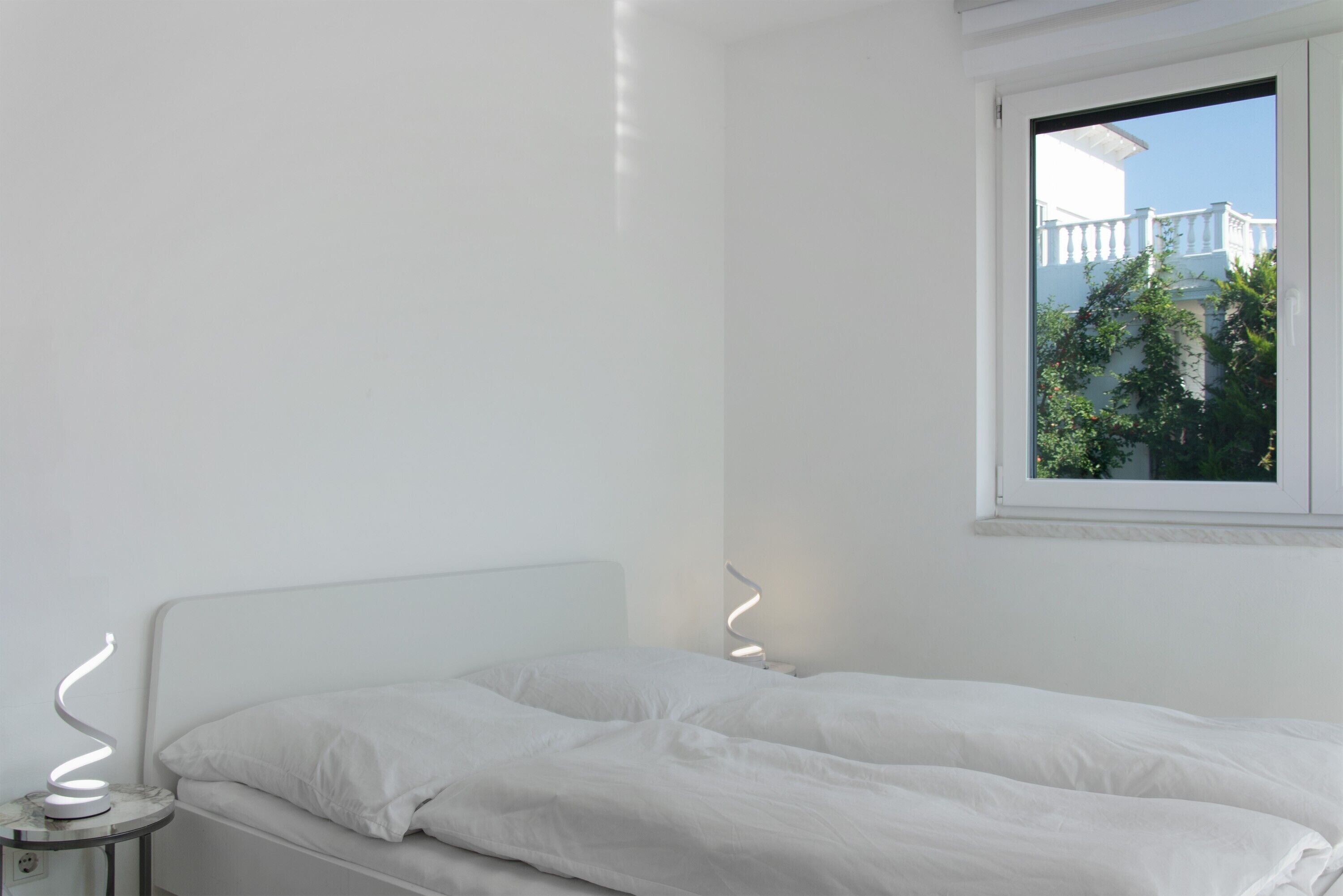 A minimalist bedroom with a white bed, a small bedside table, a modern lamp, and a window with a view of a white building and greenery.