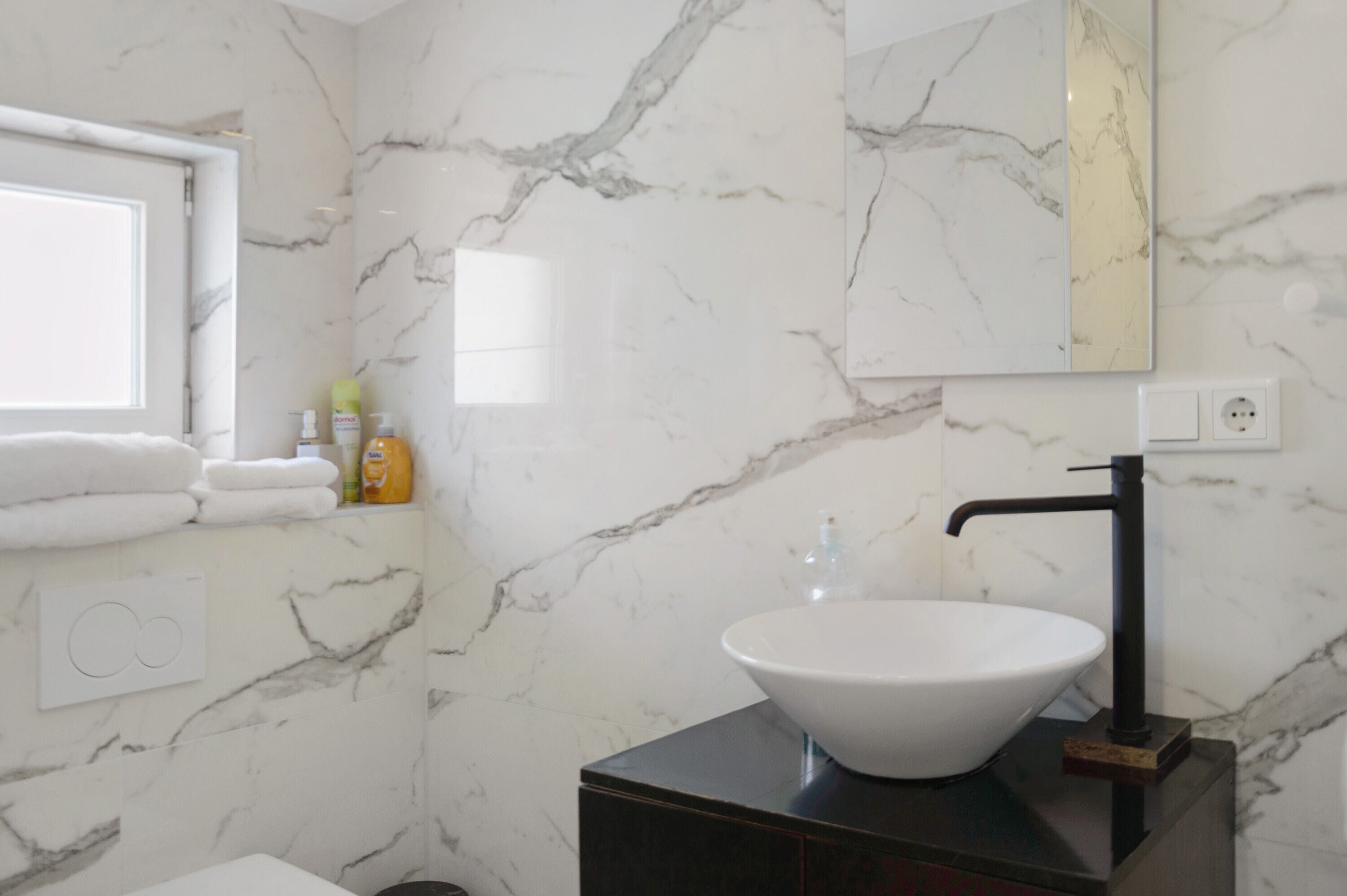 A bathroom with a marble wall, a white sink, a black faucet, and a window with towels.
