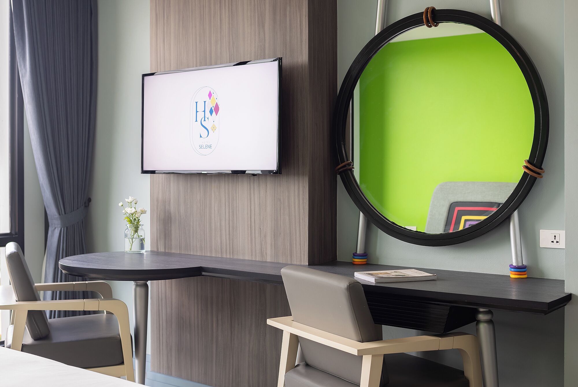 A hotel room with a desk, two chairs, a TV mounted on the wall, and a large mirror with a green frame.