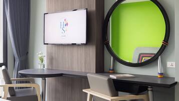 A hotel room with a desk, two chairs, a TV mounted on the wall, and a large mirror with a green frame.
