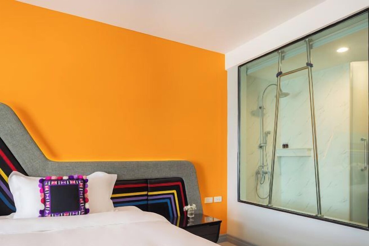 Habitación de hotel con una pared naranja, una cama con cabecera colorida y un ventanal con zona de ducha.