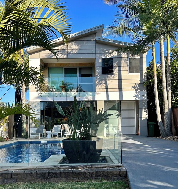 A modern two-story house with a glass front, a swimming pool, and palm trees. - The Palm - Beachside Home with Heated Pool, Cinema & Games Room (Mudjimba)
