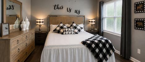 A bedroom with a bed, dresser, nightstands, lamps, a ceiling fan, a window with curtains, and wall decor.