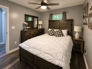 Un dormitorio con cama, cómoda, mesita de noche, ventilador de techo y ventana con cortinas.