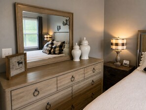 Een slaapkamer met een houten dressoir, een grote spiegel en een nachtkastje met een lamp.