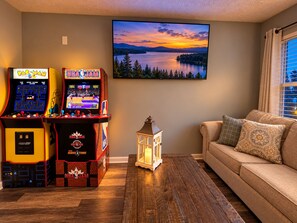 Un salon avec deux bornes d’arcade, un téléviseur diffusant une magnifique image de coucher de soleil et un canapé confortable.