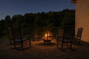 A fire pit with four wooden chairs arranged around it, set on a gravel surface with a forested hill in the background. - Amazing Beds!-2 miles to Dollywood-The Island-Tanger (Sevierville)