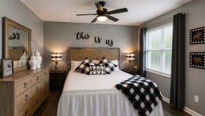 Un dormitorio con cama, cómoda, mesitas de noche, lámparas, ventilador de techo, ventana con cortinas y decoración en las paredes.