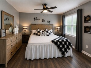 A bedroom with a bed, dresser, nightstands, lamps, a ceiling fan, a window with curtains, and wall decor.