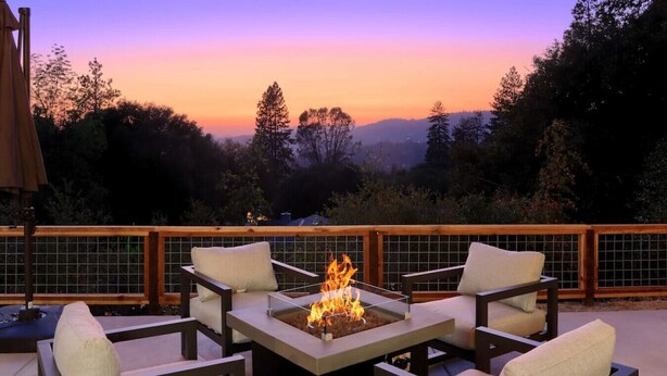 A deck with a fire pit and four chairs, surrounded by a wooden railing, overlooking a forested landscape. - Hot Tub, Fire Pit, Privacy, Game Room, and Amazing Mountain Sunset Views! (Oakhurst)