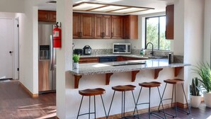 A modern kitchen with wooden cabinets, a granite countertop, a built-in microwave, and a stainless steel refrigerator. There are four bar stools and a potted plant. - Hot Tub, Fire Pit, Privacy, Game Room, and Amazing Mountain Sunset Views! (Oakhurst)
