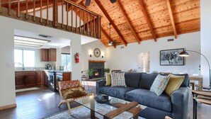 A spacious living area with a high ceiling, a fireplace, and a modern kitchen in the background. - Hot Tub, Fire Pit, Privacy, Game Room, and Amazing Mountain Sunset Views! (Oakhurst)