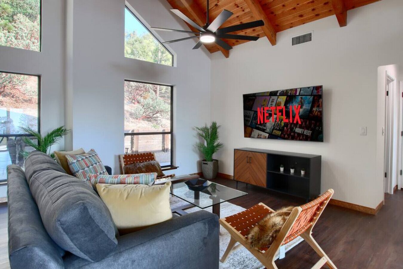 Ein geräumiges Wohnzimmer mit einem grauen Sofa, einem Glastisch und einem Holzstuhl. Ein großer an der Wand montierter Fernseher zeigt das Netflix-Logo.