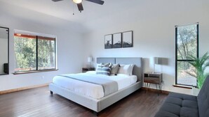 A modern bedroom with a large bed, a ceiling fan, and large windows offering a view of trees. - Hot Tub, Fire Pit, Privacy, Game Room, and Amazing Mountain Sunset Views! (Oakhurst)