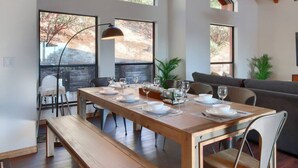 A dining area with a wooden table set for six, large windows, and a modern lamp. - Hot Tub, Fire Pit, Privacy, Game Room, and Amazing Mountain Sunset Views! (Oakhurst)