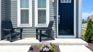Uma casa com porta frontal azul escura, duas janelas com venezianas brancas e duas cadeiras Adirondack pretas no alpendre.