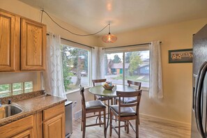 A kitchen with wooden cabinets, a granite countertop, a dining table with chairs, and a window with curtains. - Comfy & Cozy Kalispell Home: Walk to Downtown (Kalispell)