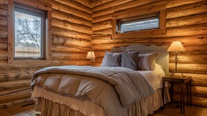 Un dormitorio tipo cabaña de madera con una cama grande, dos ventanas y una mesita de noche con lámpara.