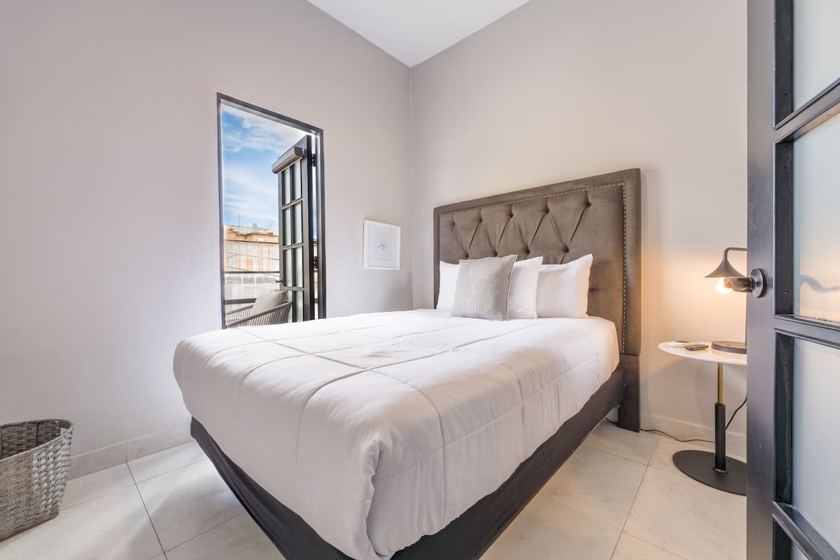 A neatly made bed with a tufted headboard, a small bedside table with a lamp, and a view of the city through a window.