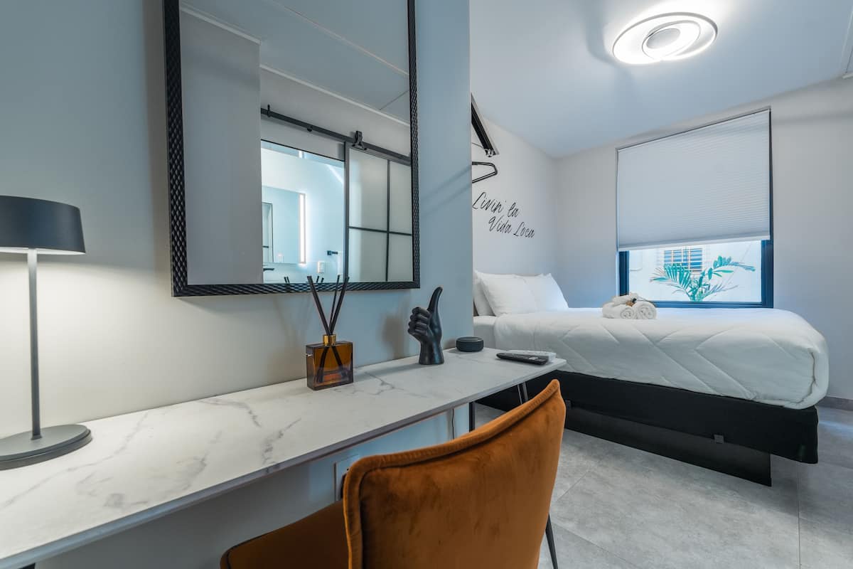 A modern hotel room with a bed, a desk with a lamp, a chair, and a large mirror.