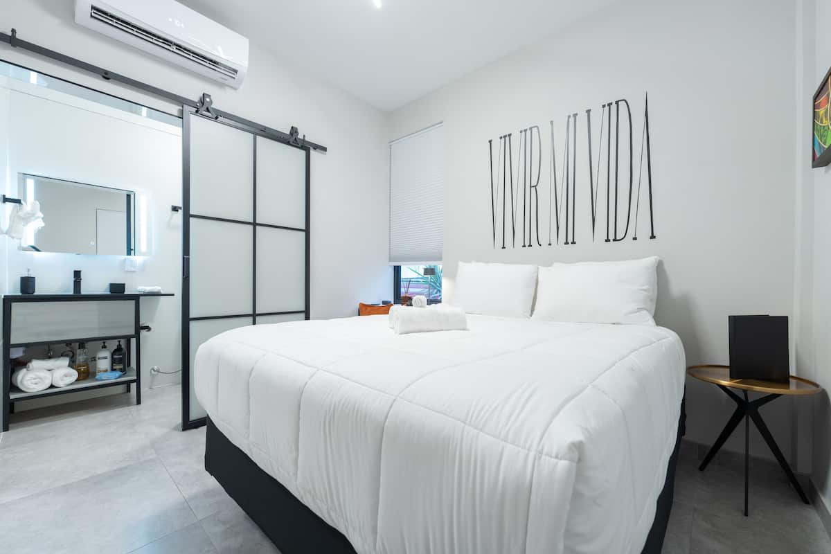 A modern hotel room with a large bed, a bedside table, a mirror, a sliding door, and a wall-mounted artwork.