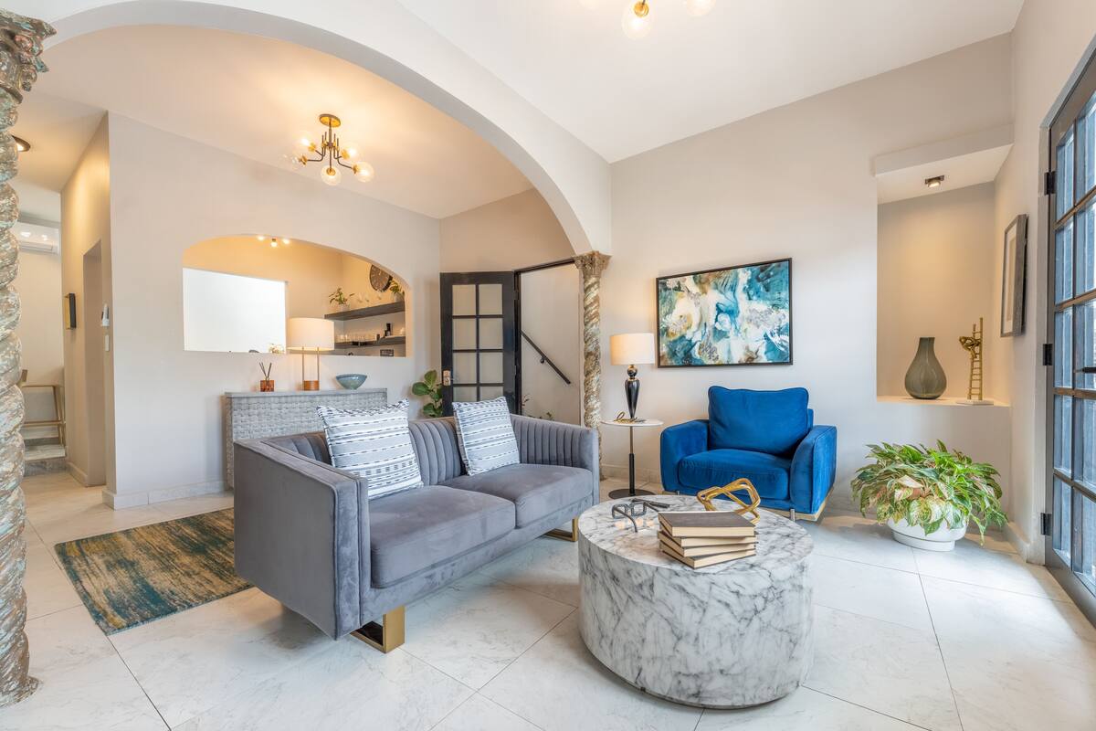 A modern living room with a grey sofa, a blue armchair, a marble coffee table, and a large window.