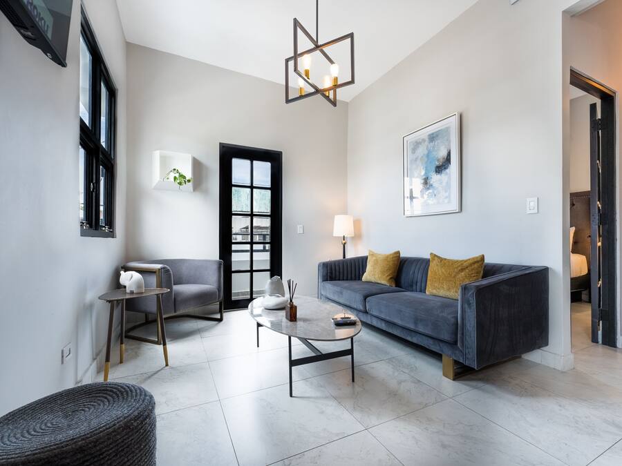 A modern living room with a grey sofa, a small round table, and a black chair. There is a door with a black frame and a window with blinds.