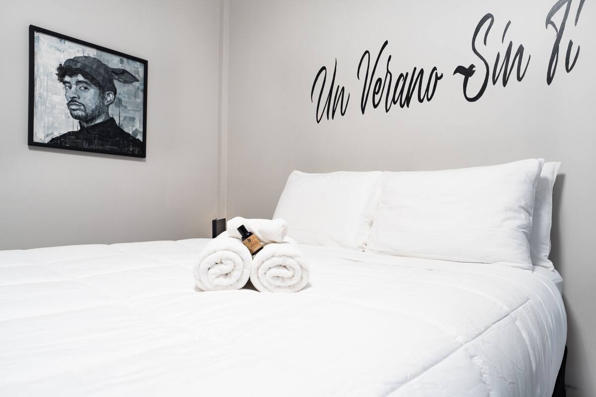 A neatly made bed with white linens and rolled towels, a framed artwork on the wall, and a wall-mounted sign with text.