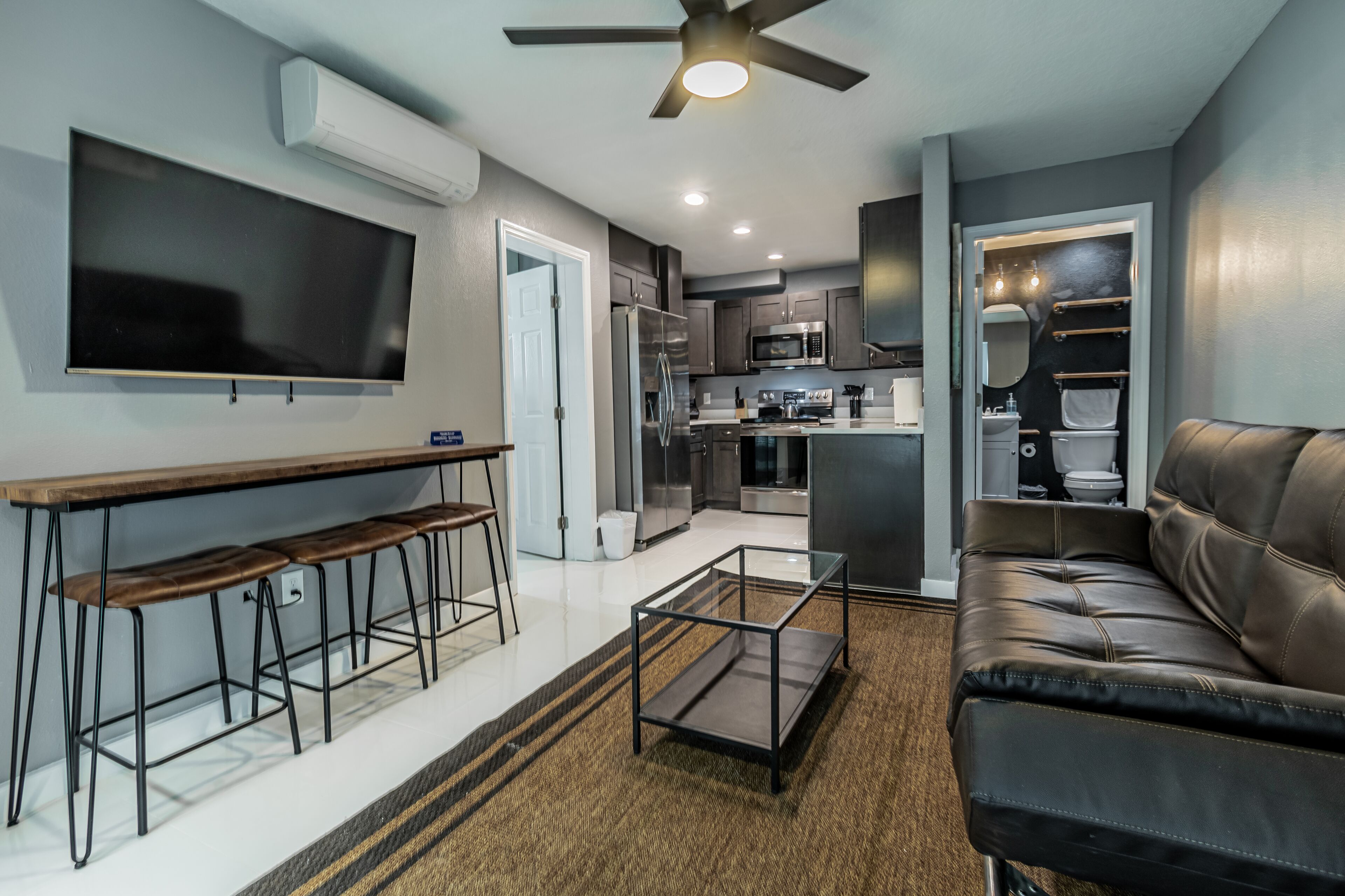 A modern living room with a sofa, a coffee table, and a TV mounted on the wall. There is a kitchen area with a refrigerator, microwave, and sink.