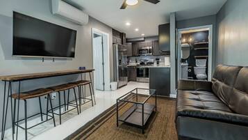 A modern living room with a sofa, a coffee table, and a TV mounted on the wall. There is a kitchen area with a refrigerator, microwave, and sink.