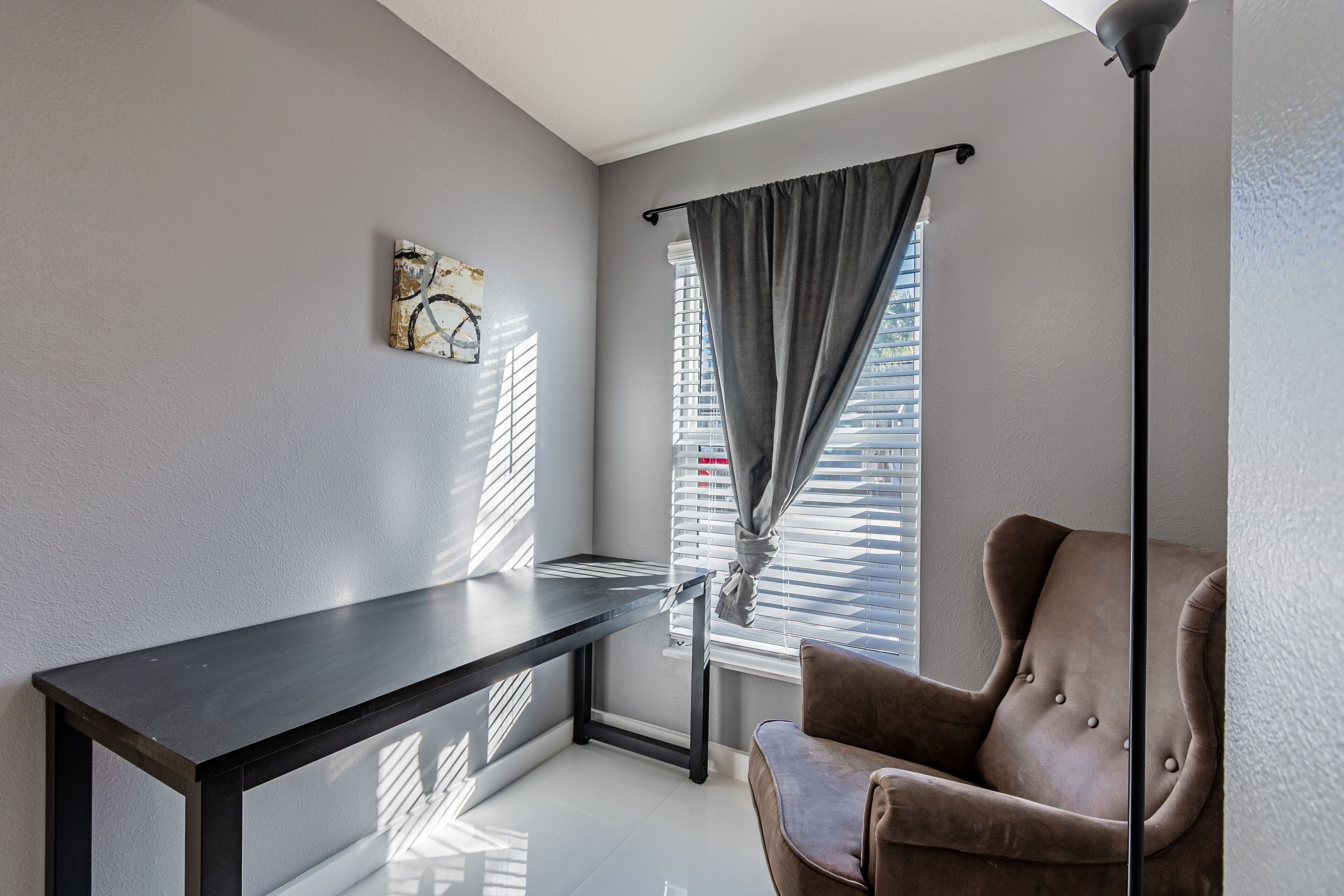 A room with a dark wooden desk, a brown leather chair, a window with blinds, and a wall-mounted artwork.