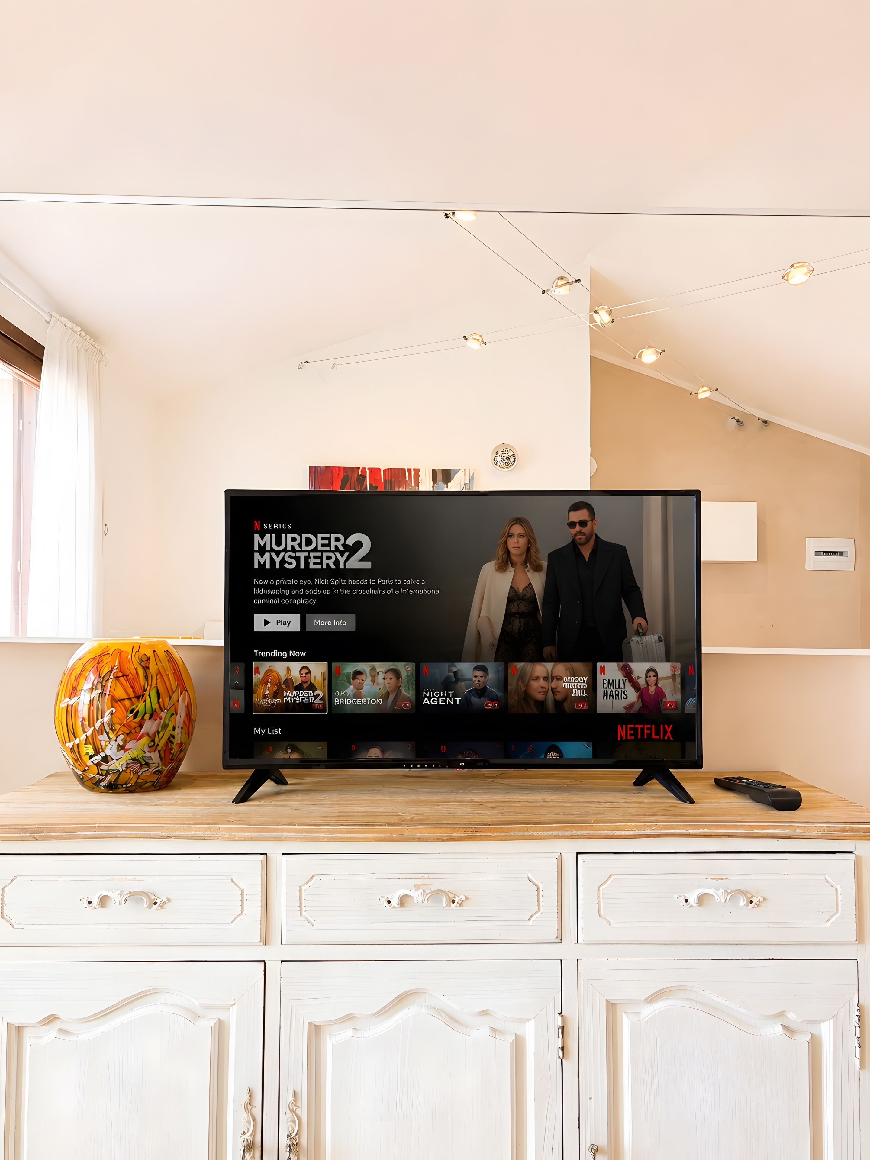 Ein Fernseher zeigt 'Murder Mystery 2' auf Netflix, mit einer Vase und einer Fernbedienung auf dem Standfuß.