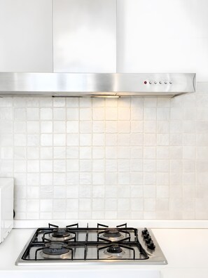 Une cuisine équipée d’une hotte en acier inoxydable, d’une cuisinière à gaz à quatre brûleurs et de carreaux de céramique blancs disposés en motif métro sur le mur.