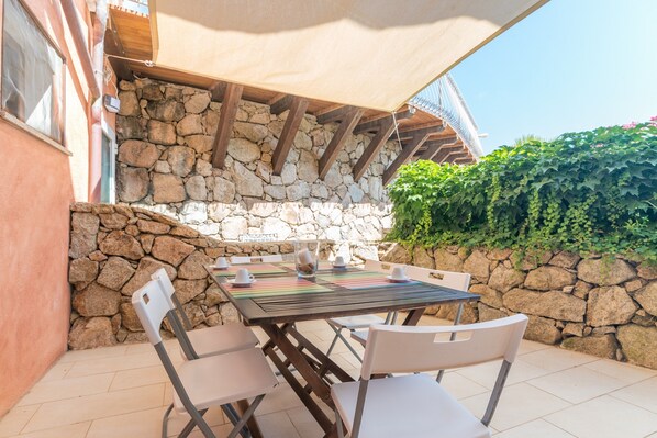 An outdoor patio with a stone wall, a wooden table, white chairs, and a shaded area.