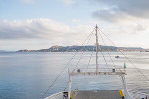 Un barco con mástil y aparejo, agua tranquila, tierra a lo lejos con edificios y un cielo parcialmente nublado.